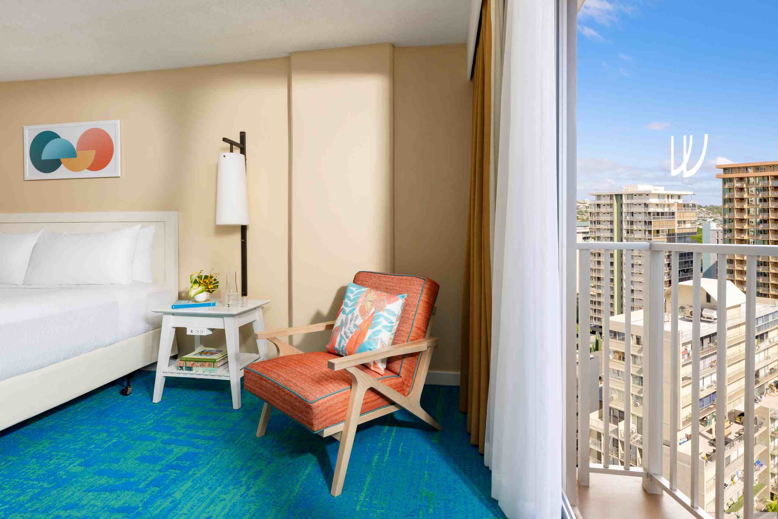 A hotel room with a partial ocean view, featuring a bed, orange chair, white table, and balcony with a city view.