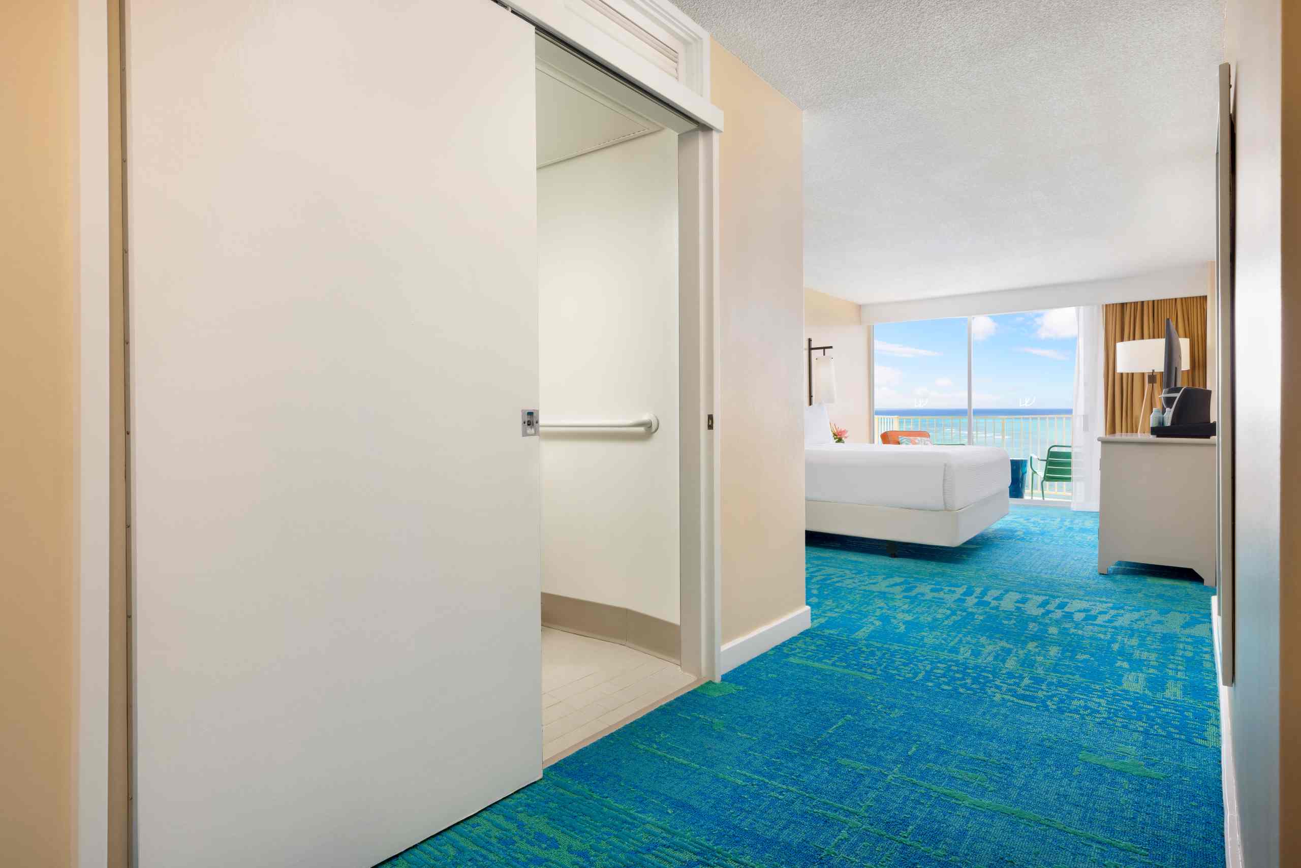 A wide view of a hotel room with an ocean view, showing a king-sized bed, a balcony, and a blue carpeted floor.