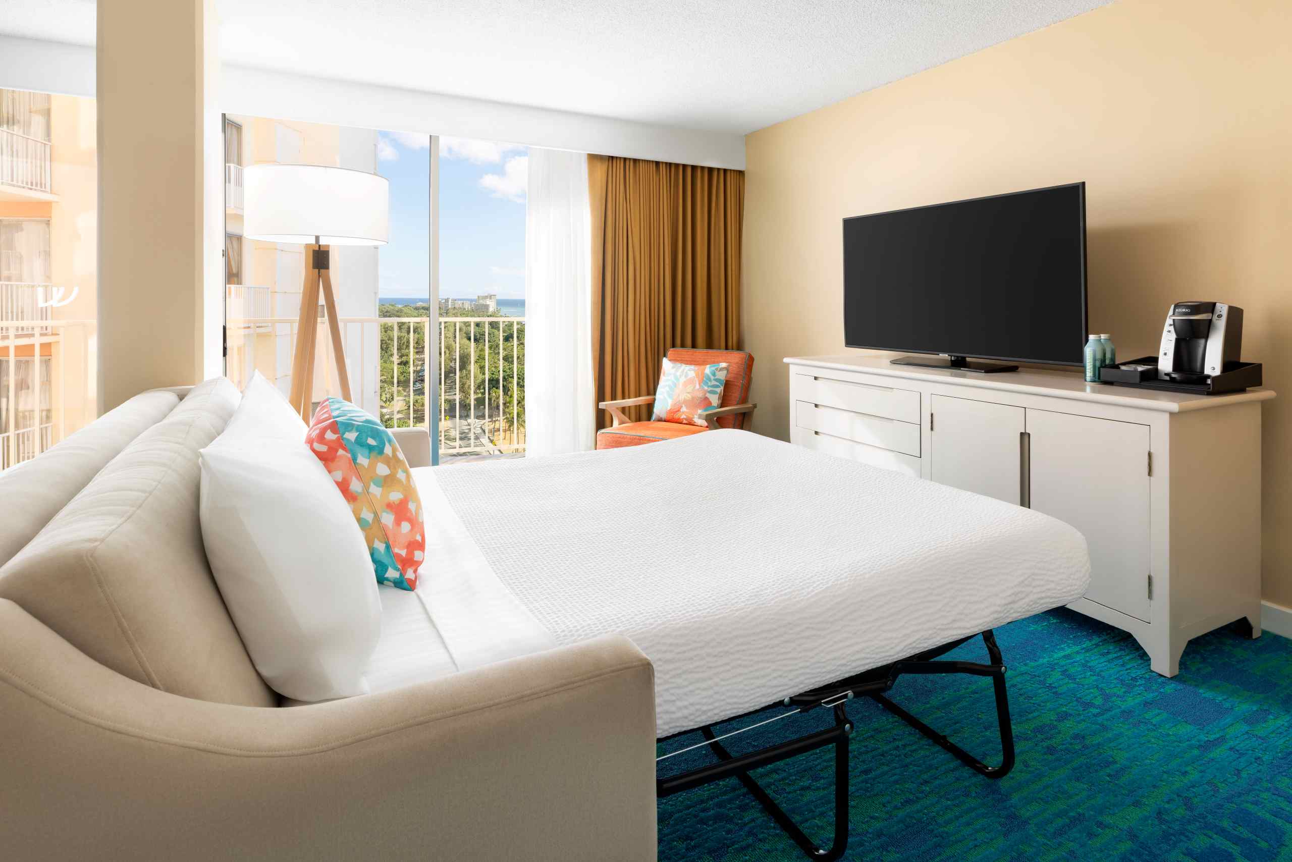 A hotel room with a sofa bed, TV, coffee maker, and balcony with ocean view