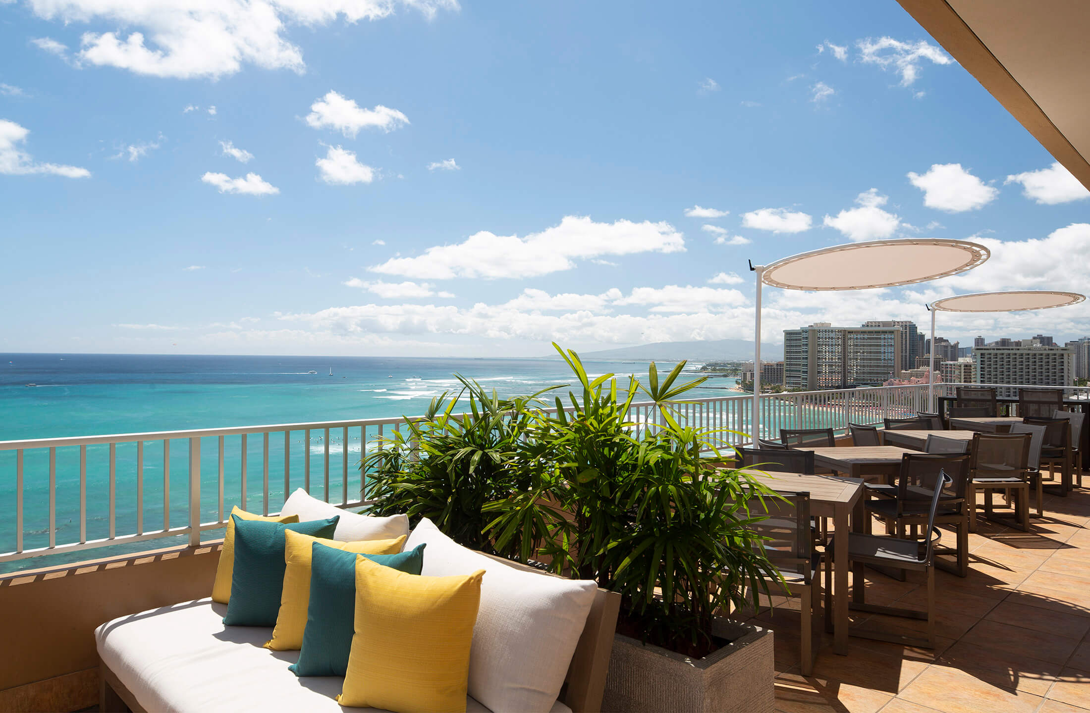 A balcony with a view of the ocean and city skyline, featuring a couch, pillows, plants, and tables with chairs.
