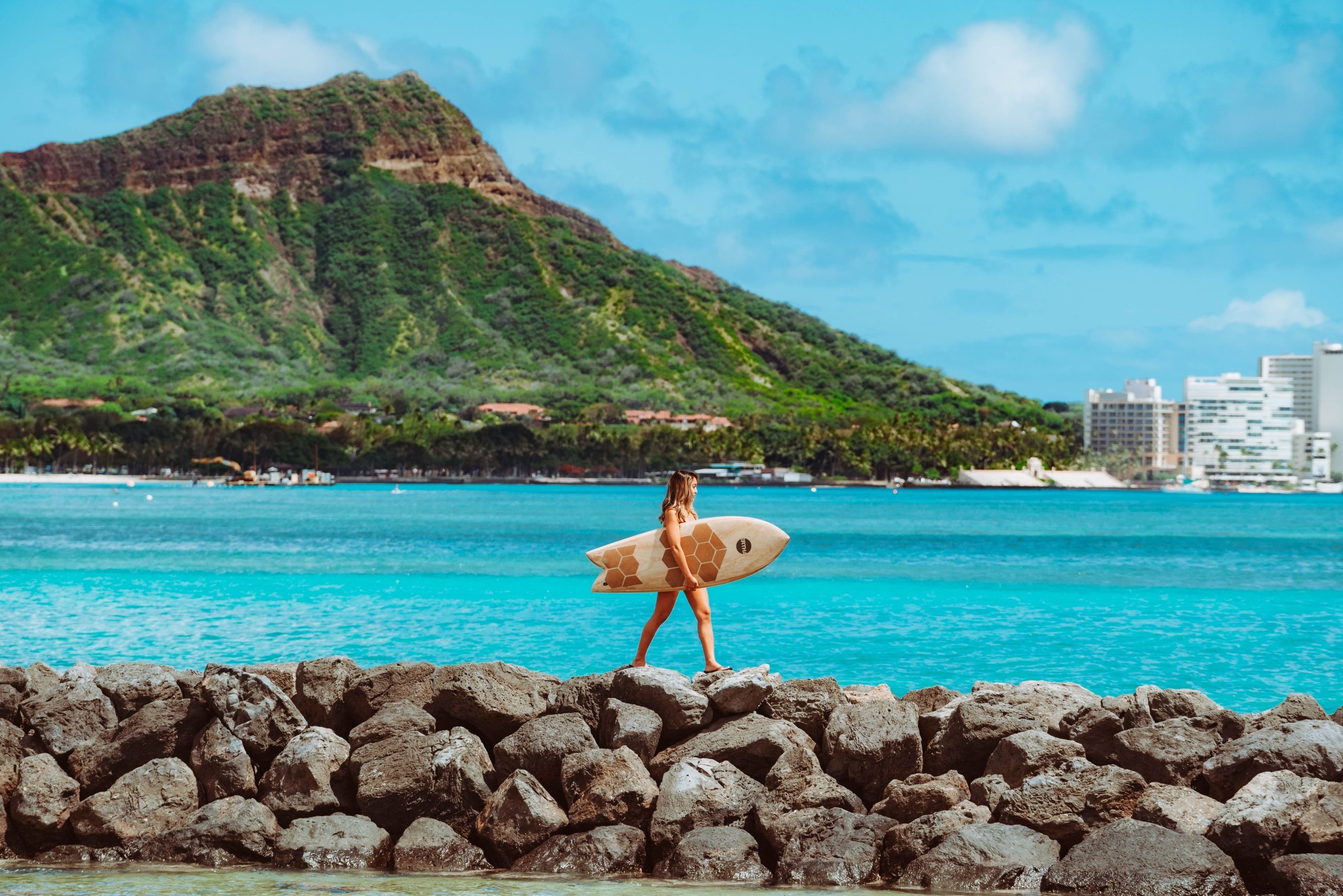 Woman walking on rocks, holding a surfboard, with Hawaiian beach in the background during the day