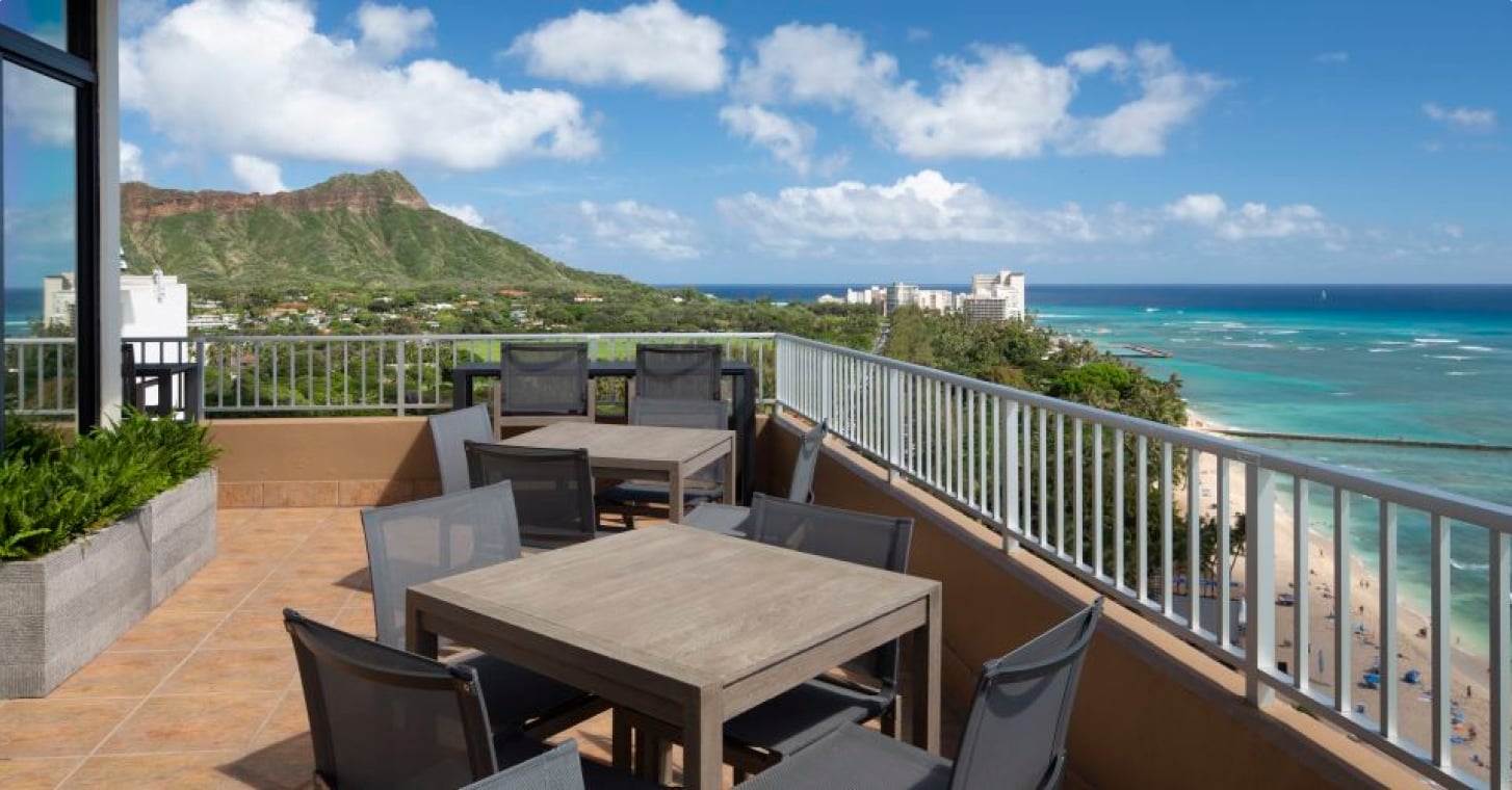 A balcony with a dining area overlooking the ocean and a mountain.