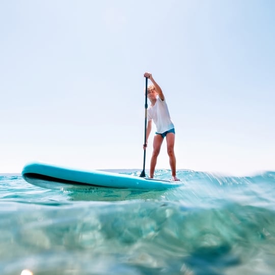 Under the water view angle to the smiling blonde teenager boy rowing stand up paddle board.