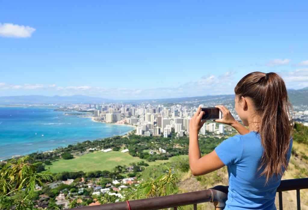 Hawaii tourist taking photo of Honolulu and Waikiki beach using smartphone camera. Woman tourist on hike visiting famous viewpoint lookout in Diamond Head State Monument and park, Oahu, Hawaii, USA.