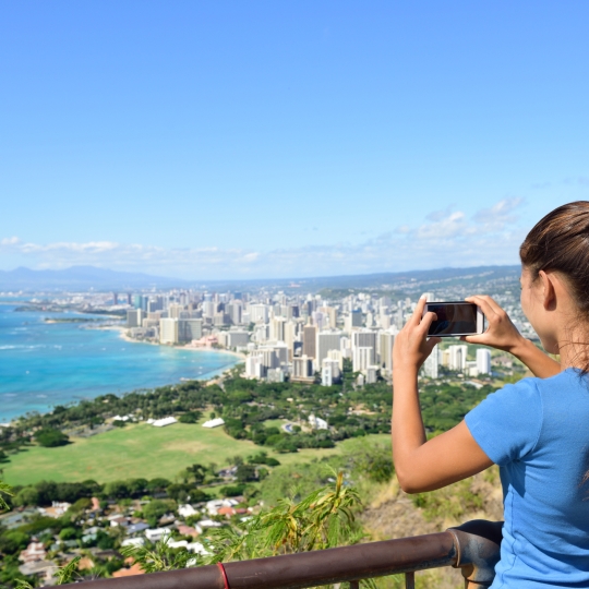 Hawaii tourist taking photo of Honolulu and Waikiki beach using smartphone camera. Woman tourist on hike visiting famous viewpoint lookout in Diamond Head State Monument and park, Oahu, Hawaii, USA.