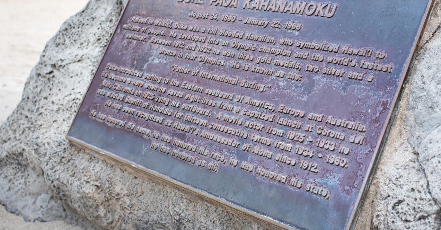 Waikiki; view of a plaque affixed to a stone, featuring inscriptions, set against a backdrop of earthy textures.