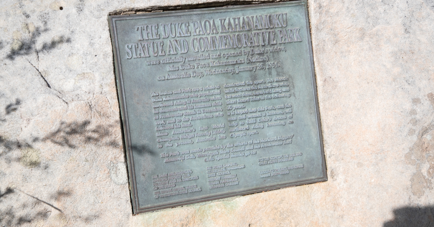Manly Beach close-up of a plaque mounted on a rugged rock surface, displaying engraved text and surrounded by natural scenery.