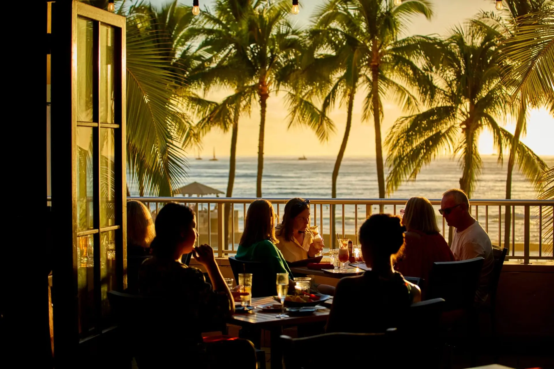 A group of people dining at a table with a view of the ocean and palm trees at sunset.